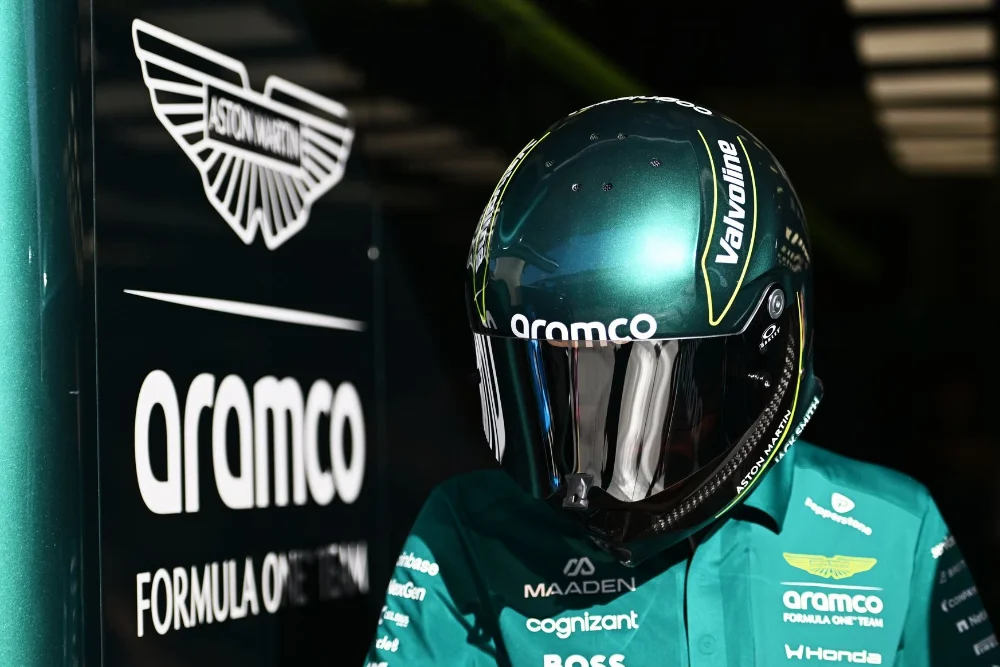 f1-2026-aston-martin-logo Gros plan sur le casque vert émeraude d'un pilote Aston Martin devant le logo ailé de la marque et le sponsor Aramco dans le garage