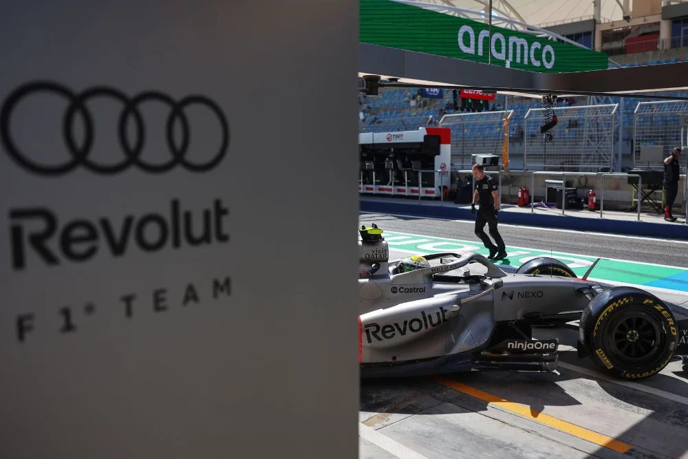 f1-2026-audi-logo Panneau officiel Audi F1 Team avec logos Revolut et les quatre anneaux, devant la monoplace dans la pitlane de Bahreïn.